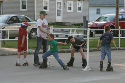 Four children and one adult rollerblading outside near houses and parked cars, with one child leaning forward.